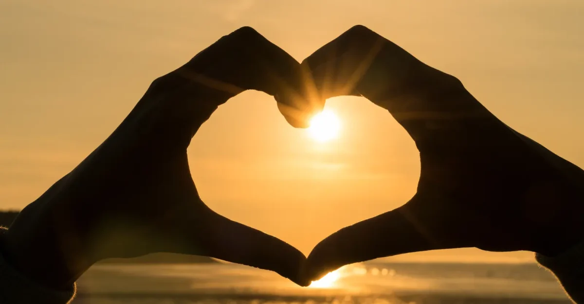 person making heart with hands in front of gold sunset